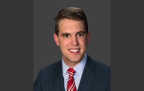 A man in a blue suit and a red and white patterned tie smiles at the camera against a gray background.