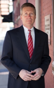 A man in a suit and tie stands outside in front of a red brick building.