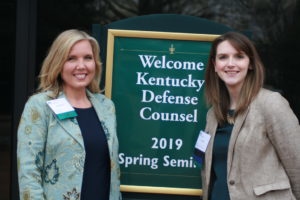 Two women stand next to a sign welcoming the Kentucky Defense Counsel 2019 Spring Seminar.