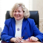 A smiling woman with blonde curly hair wearing a blue blazer sits at a desk.